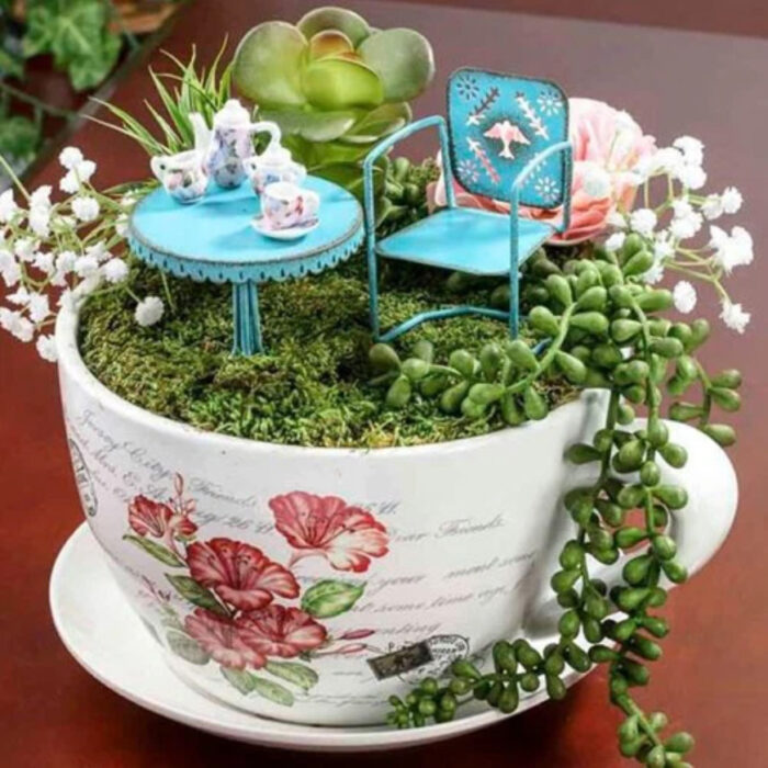 Mini jardín en taza blanca con diseño de rosas y varias suculentas, con mesa y silla de jardín azules