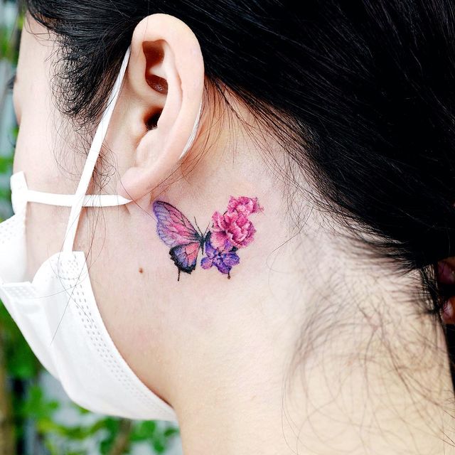 Tatuaje de mariposa detrás del oído en tonos rosas con flores desprendidas; 15 Bellos tatuajes con mariposas para iniciar una metamorfosis