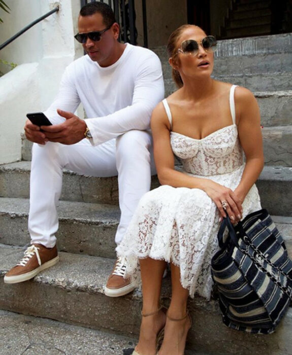 Jennifer Lopez y Alex Rodríguez sentados en unas escaleras mirando sus teléfonos