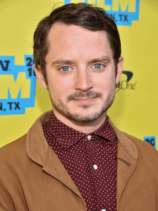 Elijah Wood usando una camisa burgundy y saco camel