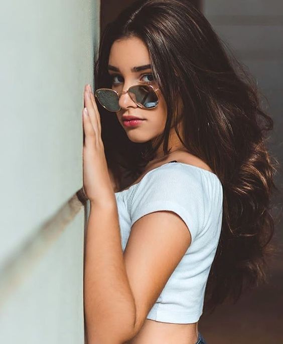 Chica morena clara posa recargando sus manos en la pared mientras viste blusa blanca y lentes de sol
