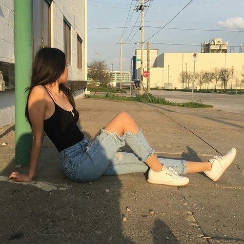 Chica delgada con blusa de tirantes negra y pantalón mezclilla posa sentada en el piso mientras se recarga en un poste verde