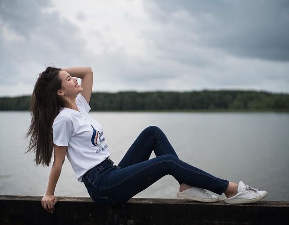 Chica sonriente con cabello largo y oscuro posa sentada con las piernas extendidas mientras mira hacia arriba y pone su mano en la cabeza