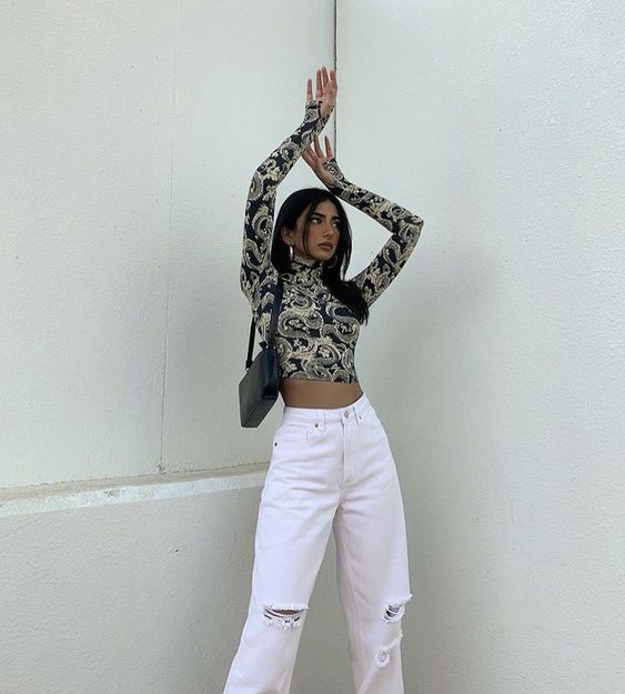 Chica morena y delgada posa con los brazos extendidos hacia arriba vistiendo pantalón blanco y blusa pegada de cuello de tortuga