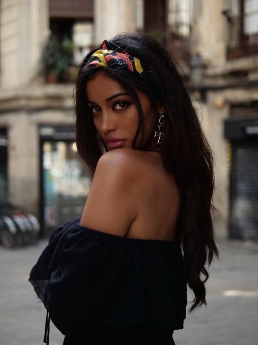 Chica morena con cabello largo y negro y una diadema amarilla con rojo y azul en la cabeza, viste una blusa al hombro color negro