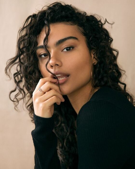 Chica morena de cabello rizado vistiendo blusa negra de manga larga con grandes arracadas y posando con su mano derecha en sus labios