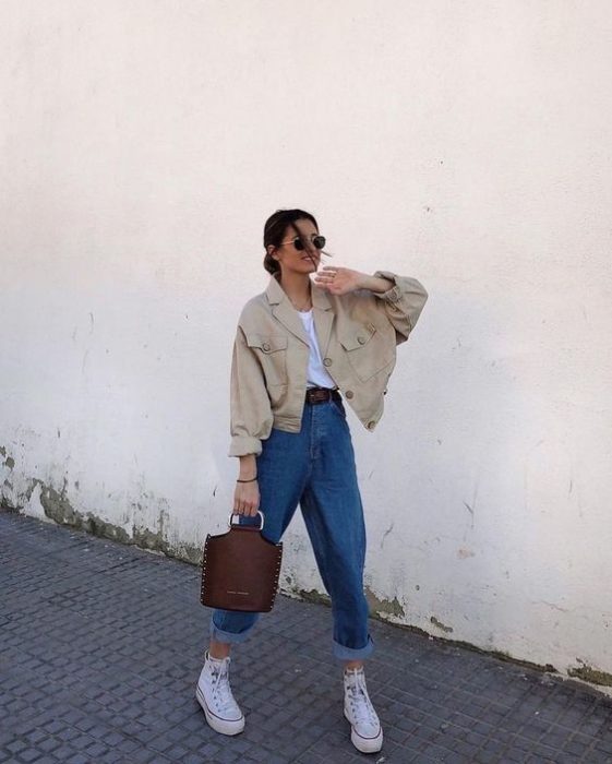 chica de cabello castaño usando lentes de sol, cazadora beige, playera blanca, bolso café de piel, jeans slouchy y tenis deportivos blancos