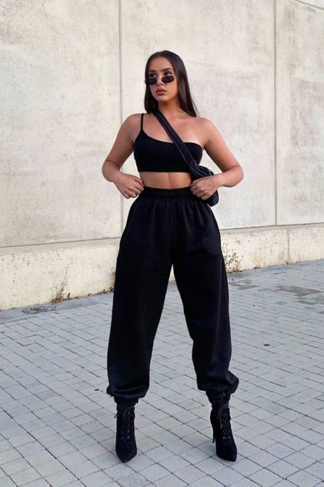 chica de cabello castaño largo usando un top negro, pants deportivos negros, botas de tacón y bolsa negra crossover