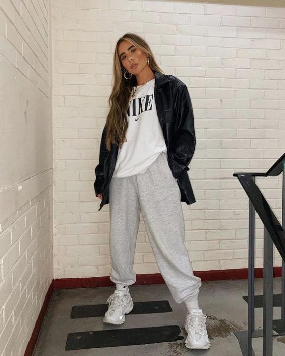chica de cabello teñido rubio usando chaqueta larga de piel negro, camiseta blanca, pants grises y tenis blancos deportivos