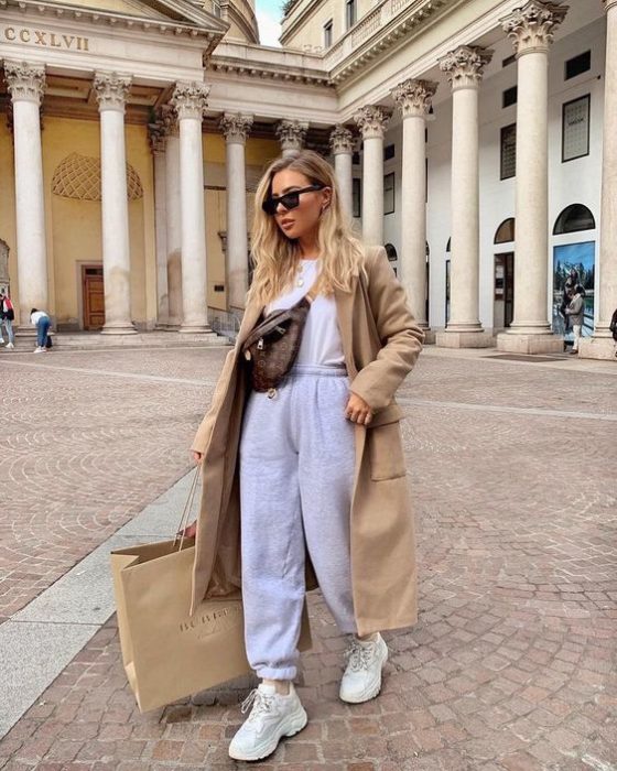 chica rubia con lentes de sol, haciendo compras, usando una bolsa tipo crossover de piel, camiseta blanca, abrigo beige, pants grises y tenis blancos deportivos