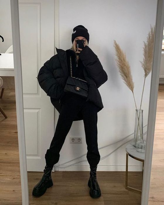 chica rubia tomándose un selfie con un beanie negro, chamarra oversized negra, top negro, bolso de piel negro, pantalones pants negros y botas de cuero