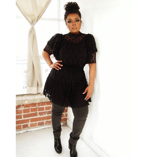 Yvette Nicole Brown posando de pie con una blusa larga negra, pantalón gris y botines negros altos