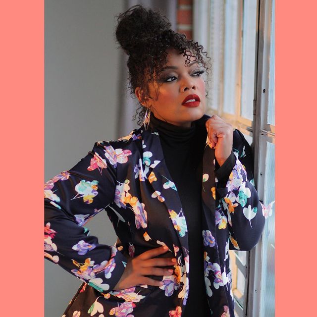 Yvette Nicole Brown posando recargada en una ventana vistiendo una blusa negra de cuello alto con un saco floreado
