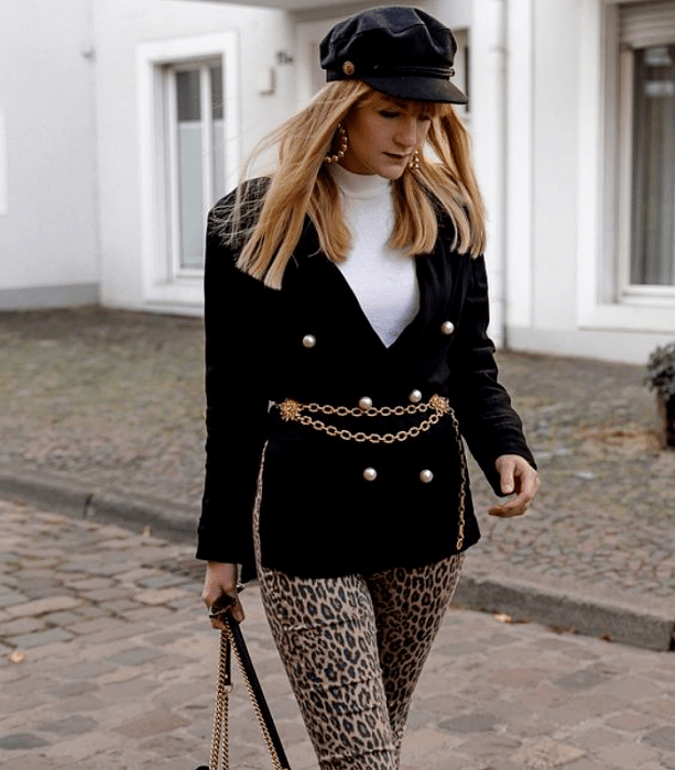 chica rubia con sombrero negro, saco negro con cinturón dorado metálico, top blanco de cuello alto, pantalón de animal print café