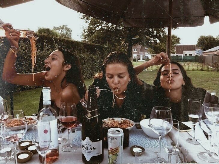 Adolescentes comiendo pasta con las manos en una mesa de mantel blanco con copas de vino y platos hondos blancos