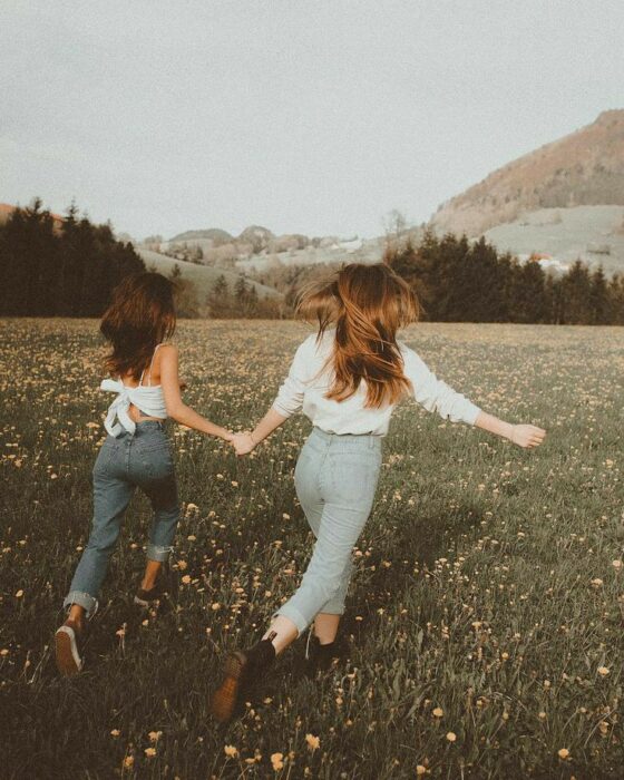 Chica de cabello castaño y chica rubia corriendo por un campo lleno de flores y una montaña al fondo