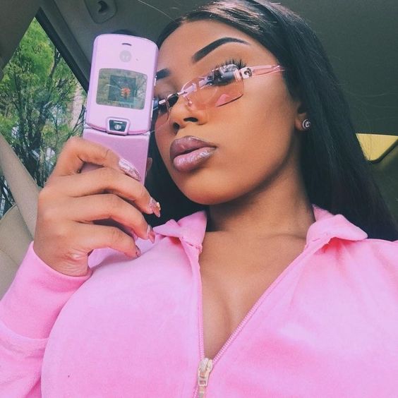 Chica morena con outfit y lentes rosas tomándose una selfie con un celular rosa y uñas largas