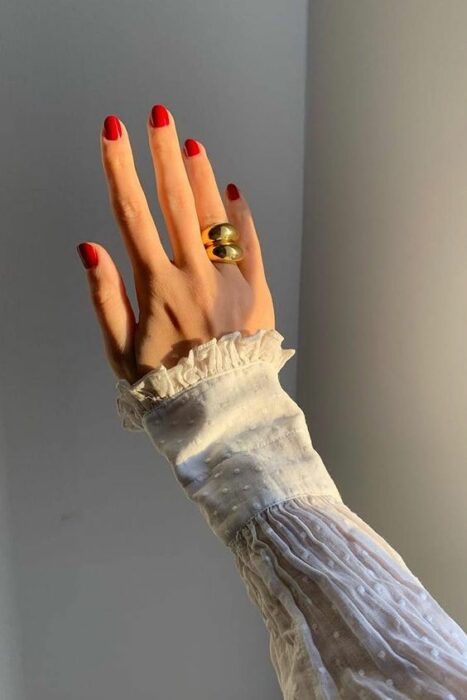 Mano de una mujer posando en el sol con las uñas pintadas de rojo y un anillo grande dorado