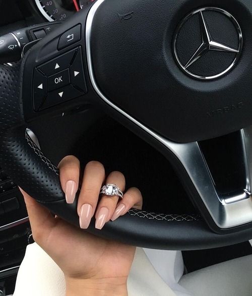 Mano de mujer con uñas acrílicas color rosa con un anillo en el dedo anular y sujetando el volante de un auto