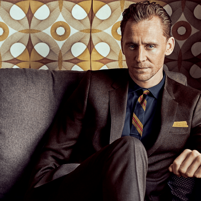 tom hiddleston con traje café oscuro, camisa azul de vestir y corbata azul con amarillo oscuro