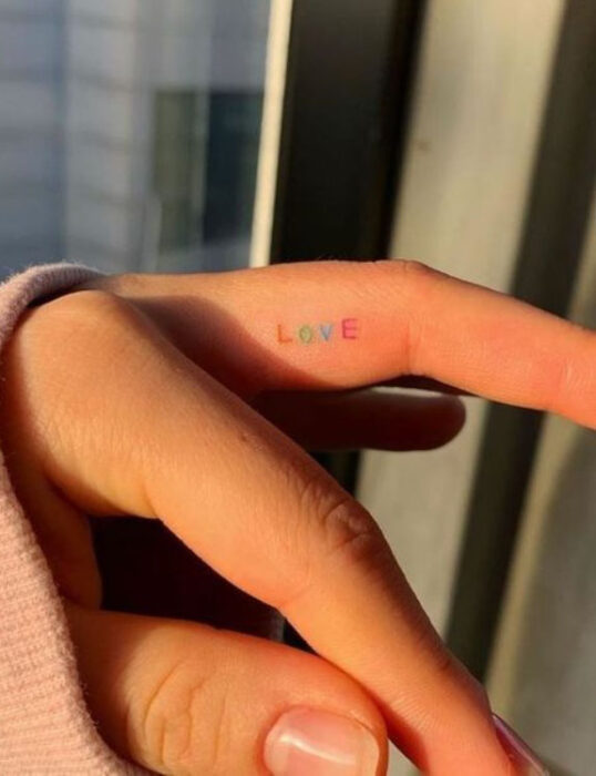 Tatuaje pequeño en la parte interna del dedo con letras de colores