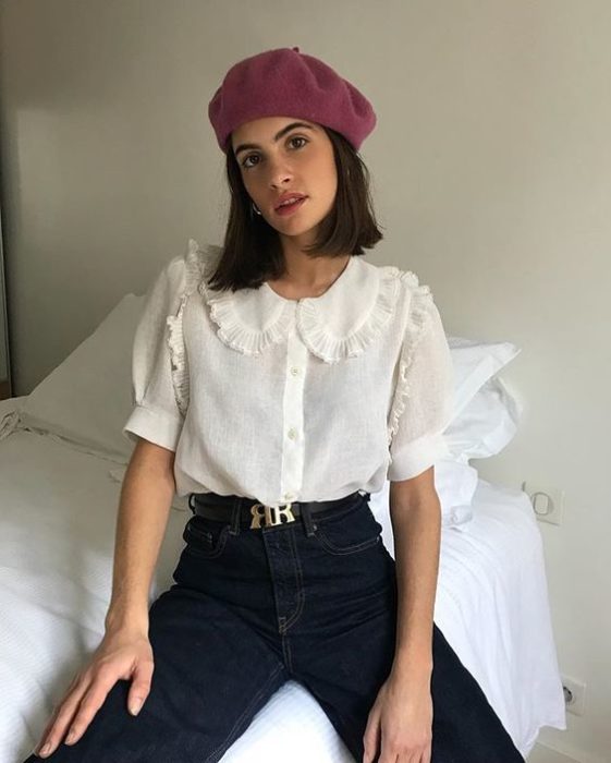 Chica delgada morena de cabello corto con gorro rosa, pantalón negro y blusa blanca con cuello bobo