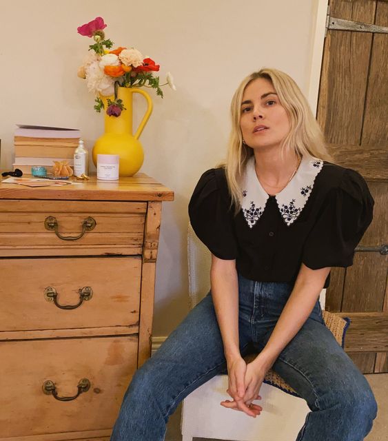 Chica rubia sentada con pantalón de mezclilla, blusa negra y cuello bobo blanco con aplicaciones de piedras