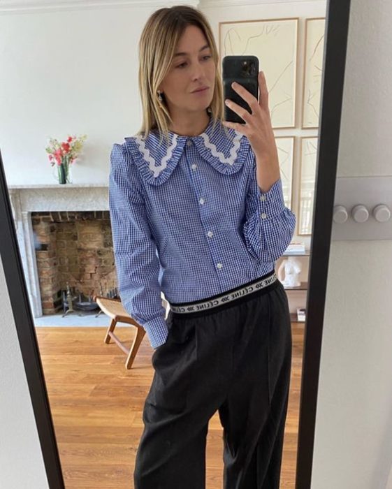 Chica rubia tomando una selfie en el espejo con pantalón negro y camisa azul con cuello bobo con perlas