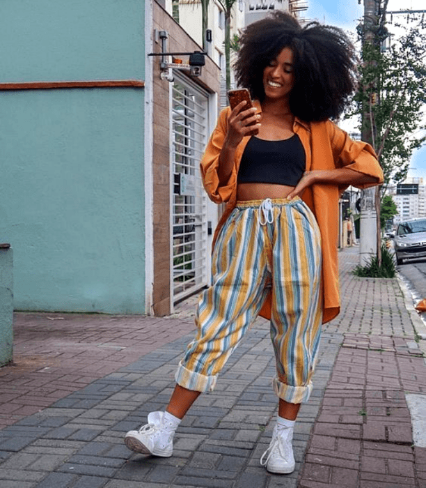 chica morena usando una camisa naranja con un crop top negro debajo y pantalones de rayas amarillos con azul y blanco y tenis deportivos blancos