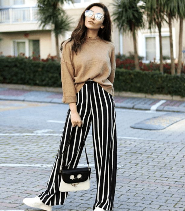 chica de cabello castaño usando lentes de sol, suéter beige, pantalón negro con rayas blancas y tenis deportivos blancos