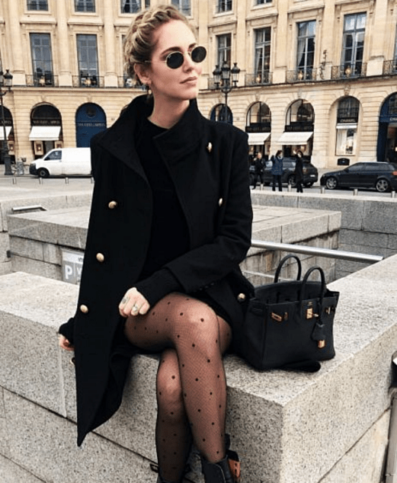 chica rubia usando lentes de sol, vestido negro, abrigo negro, bolso de mano negro pequeño, medias negras con lunares y botines