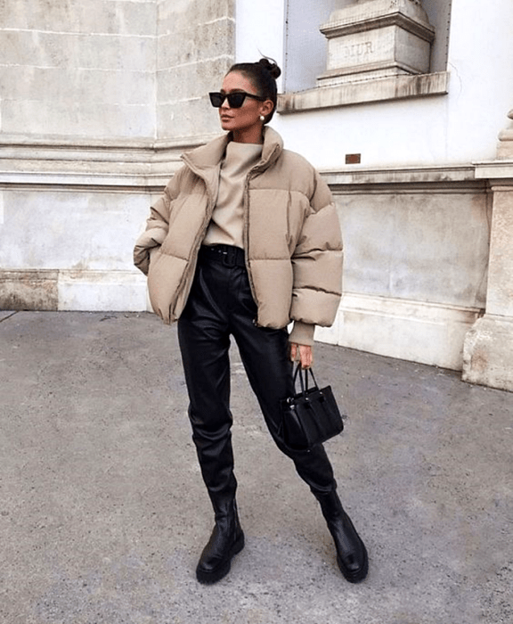 chica de cabello castaño usando lentes de sol, top beige, chamarra beige, pantalones negros de cuero a la cintura, botines negros y bolso pequeño negro