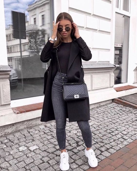 Chica de cabello largo suelto con jeans oscuros, blusa negra, saco negro y tenis blancos