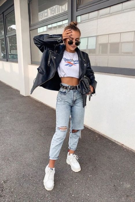 Chica rubia con cabello recogido, jeans rotos, blusa blanca y chaqueta negra de cuero