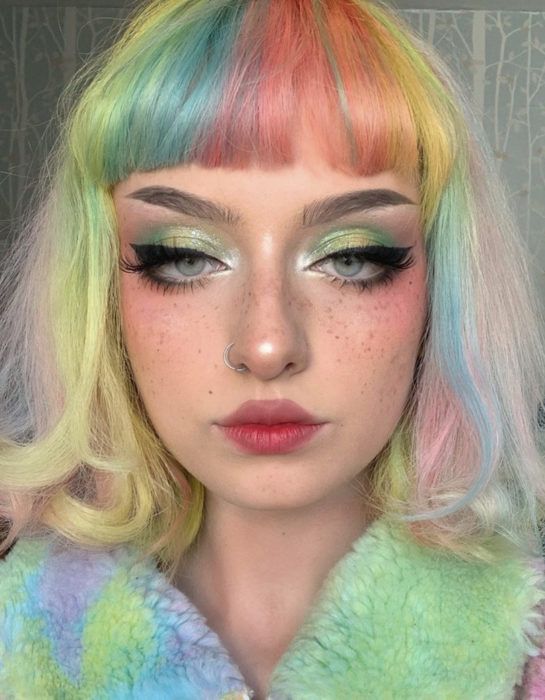 Mujer con pecas, perforación en la nariz, de ojos grises delineados, con cabello corto teñido de colores arcoíris pastel, azul, verde, amarillo, rosa y morado, ondulado, fleco