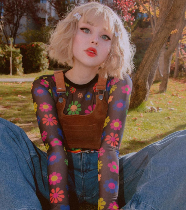 Looks aesthetic con cabello corto color rubio, ondulado, con puntas, blusa de flores hippies, overol, chica kawaii sentada en el pasto en el parque
