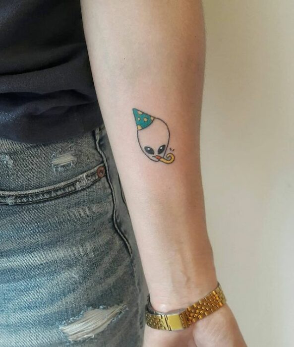 Tatuaje de marcianito con gorro de cumpleaños en el brazo
