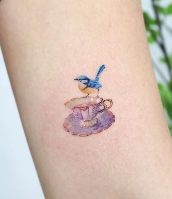 Diseños bonitos de tatuajes de acuarelas; tatuaje de pájaro azul en una taza vintage de té