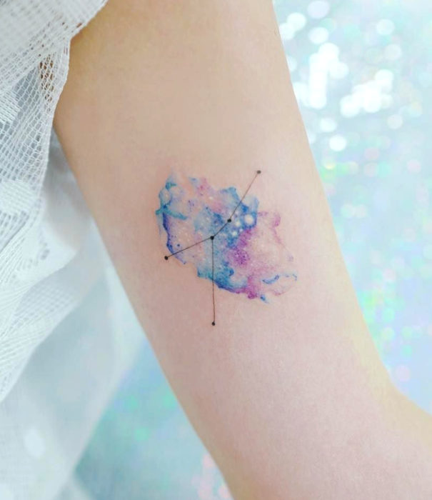 Diseños bonitos de tatuajes de acuarelas; tatuaje del universo y constelaciones en colores pastel en el brazo