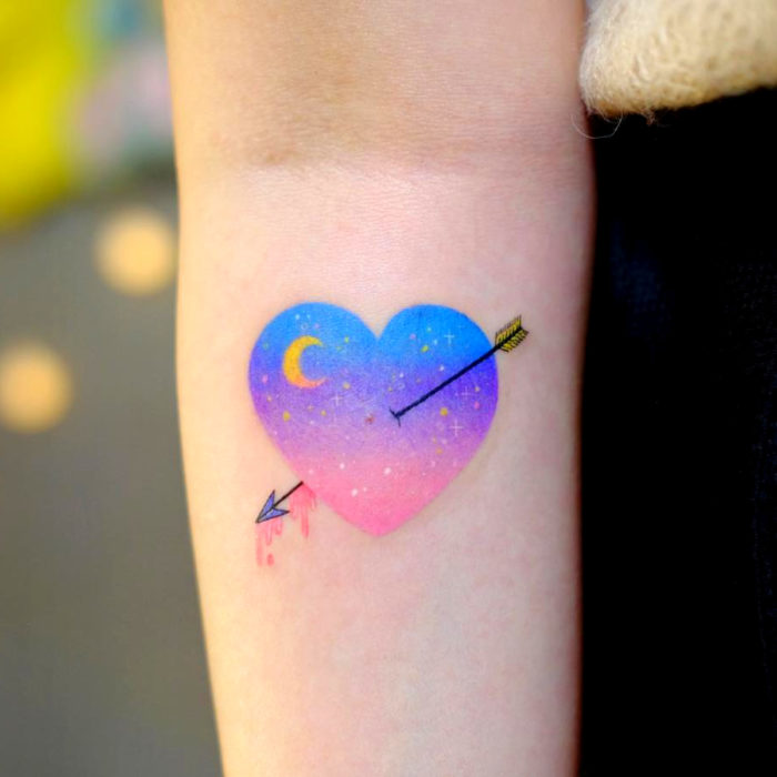 Tatuadora hace tatuajes encantadores; tatuaje de corazón con flecha y luna en el brazo