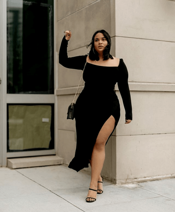 chica morena curvy usando un vestido negro ajustado de manga larga y escote cuadrado con abertura en la pierna y bolso negro pequeño