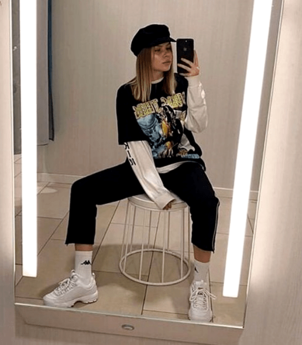 chica rubia usando un sombrero, camiseta oversized negra, top de manga larga blanco, pantalón negro, tenis blancos