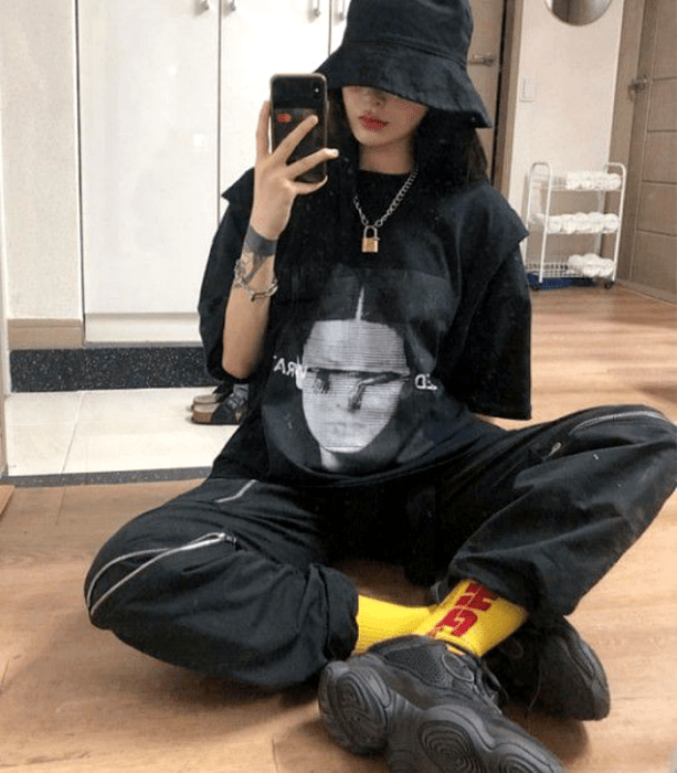chica de cabello castaño usando un bucket hat negro, camiseta negra oversized, pants negros, calcetas amarillas, tenis negros
