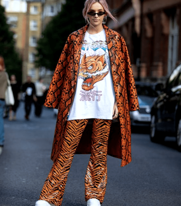 chica rubia usando lentes de sol, camiseta blanca oversized con estampado de dragón, abrigo largo de cuero naranja con líneas negras, pantalones de cuero acampanados color naranja con animal print de tigre y tenis blancos