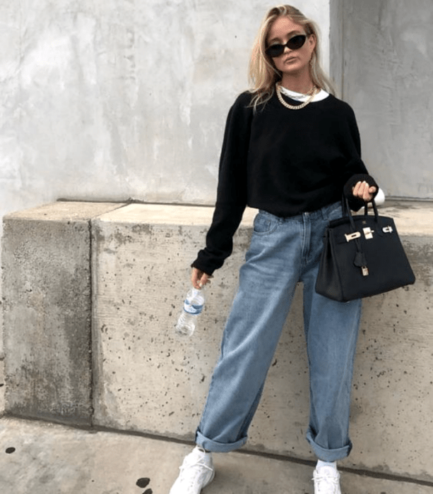 chica rubia usando lentes de sol, sudadera negra, camiseta blanca, jeans holgados y tenis blancos, bolso negro de mano