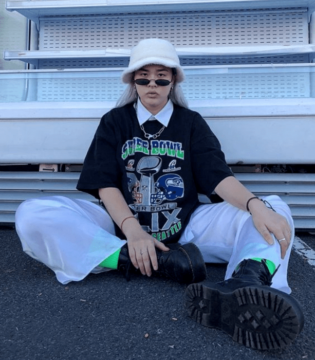 chica de cabello platinado usando un bucket hat blanco, camiseta negra oversized, camisa blanca debajo, pantalones deportivos blancos holgados y botas negras de plataforma