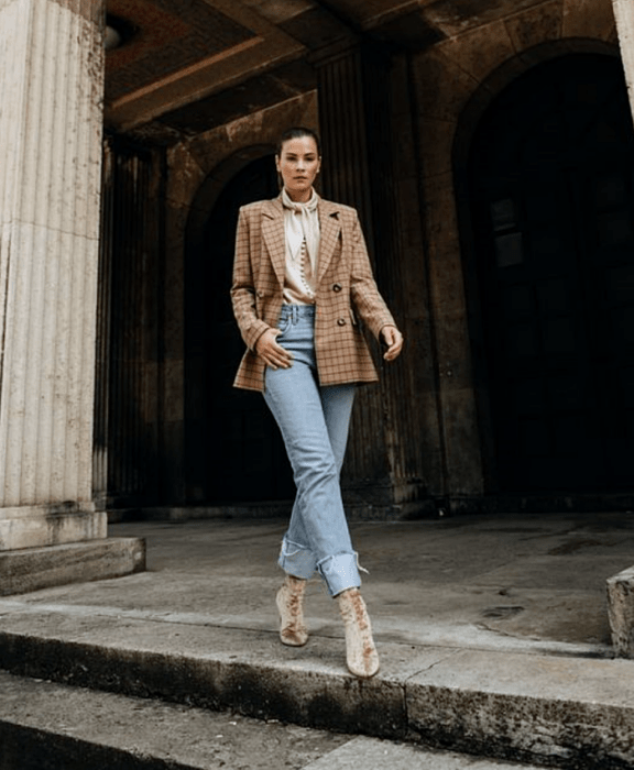 mujer con cabello castaño usando una blusa beige de satén con moño en el cuello, blazer oversized beige de cuadros, jeans a la cintura estilo mom jeans, botines estilo sock boots beige de terciopelo