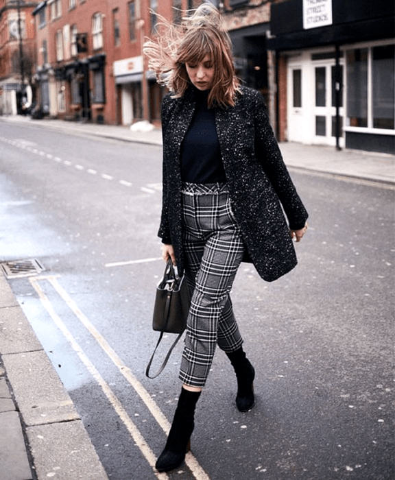 mujer con cabello claro usando un top negro, abrigo negro con lunares blancos, pantalones grises de cuadros, bolso negro de mano y botines estilo sock boot negros