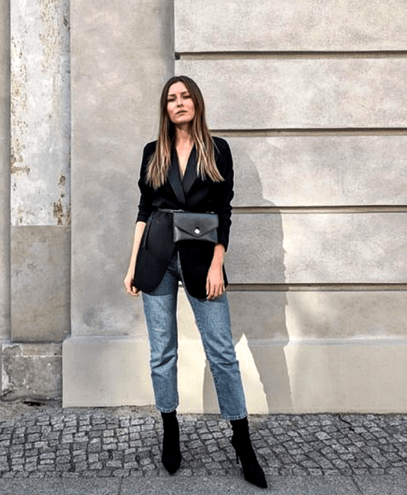 mujer con cabello claro largo usando un blazer negro oversized, bolso negro estilo cangurera, jeans rectos y botines negros estilo sock boots