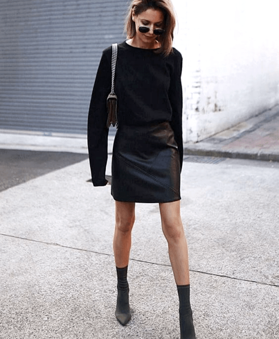 mujer con cabello castaño usando lentes de sol, suéter negro de manga larga, minifalda negra de cuero, bolso de mano negro con correa de cadena y botines estilo sock boots negros